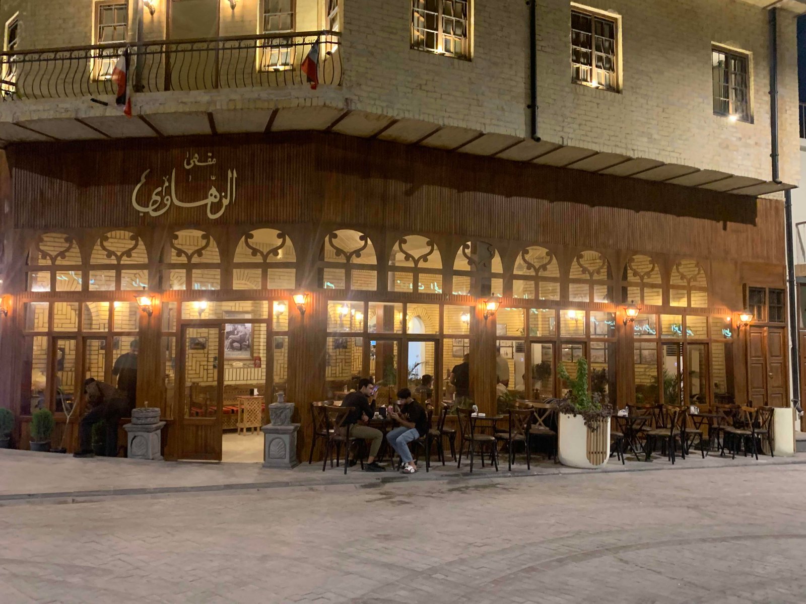 The Al-Zahawi coffeehouse in Baghdad in the evening, its wooden frontage lit, people sitting at tables on the street outside.