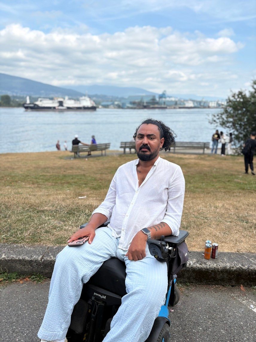 Ali Alsharahnee outdoors in his power wheelchair beside the water in Vancouver, the harbour and the North Shore mountains behind him.