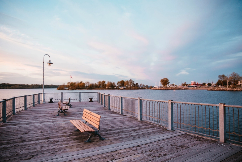Empty wooden benches on a pier beside calm water at dusk, looking out towards a far shore.